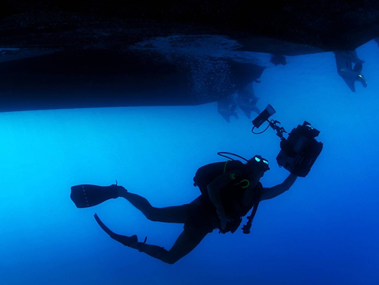 Scuba diver silhouetted against deep blue water while holding an underwater video camera near the hull of a vessel.