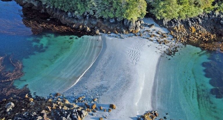 Aerial view of a small crescent shaped beach with white sand, turquoise water and rocky forested shoreline.