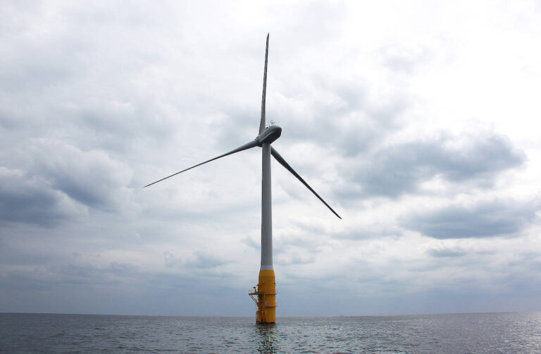 Single offshore wind turbine standing in calm open water under a cloudy sky.