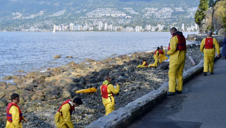 Field crew in yellow protective suits surveying an oiled shoreline along Vancouver’s Stanley Park seawall with the North Shore and Burrard Inlet in the background.