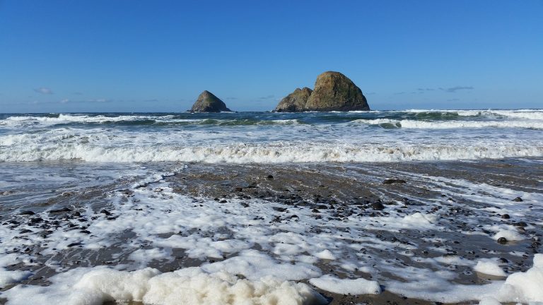 Foamy waves washing onto a sandy beach with rocky sea stacks offshore under a clear blue sky.
